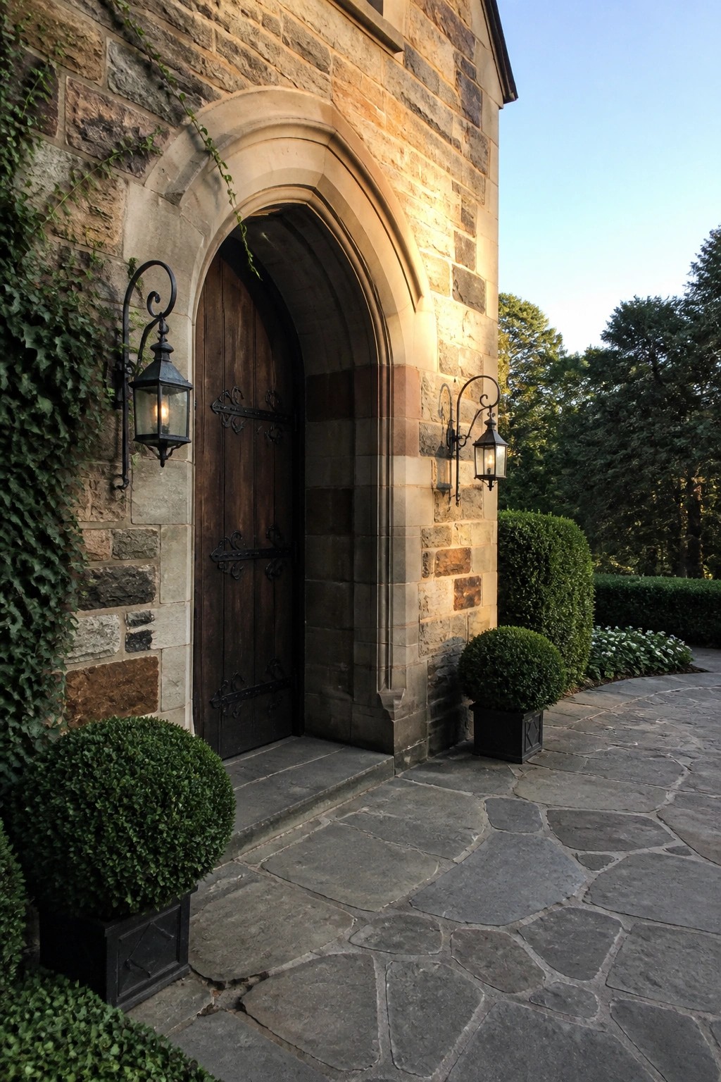 A large arched wooden door centered in a stone wall with black metal lanterns mounted on either side and boxwood shrubs in the foreground.