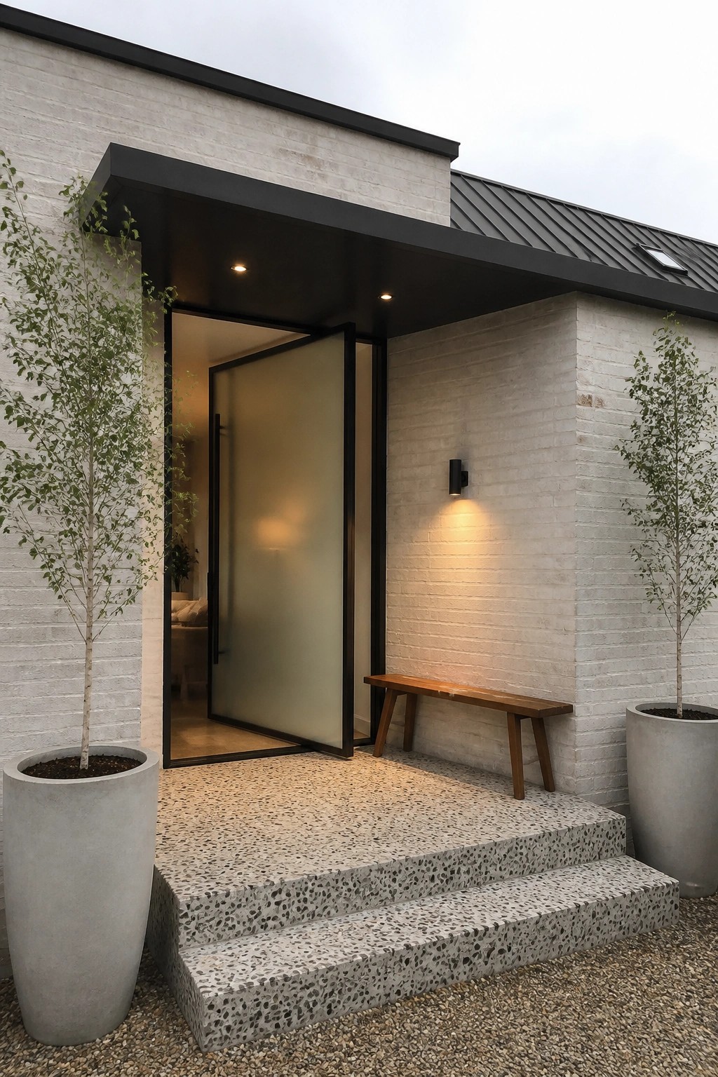 A modern white brick house exterior with a large frosted glass front door in a black frame, concrete steps, a wooden bench, wall sconce, and two large potted trees flanking the entrance.