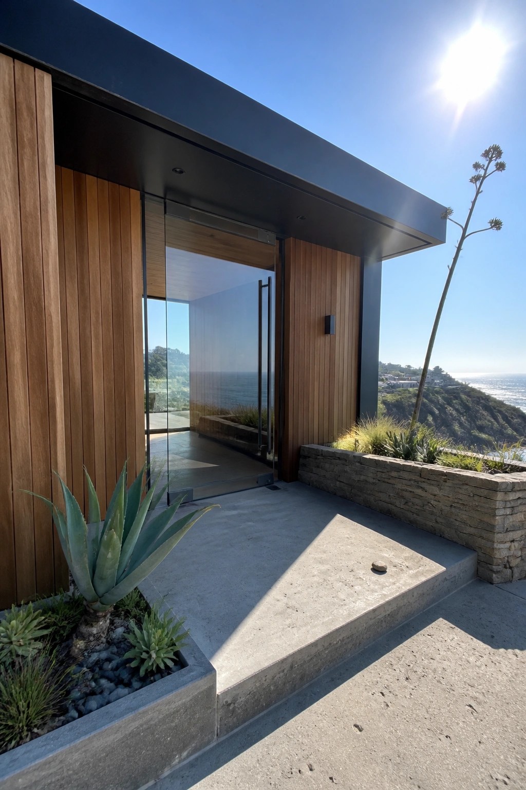 Modern house exterior with oversized glass entry doors, wood siding, concrete steps, and a stone planter beside the ocean view.