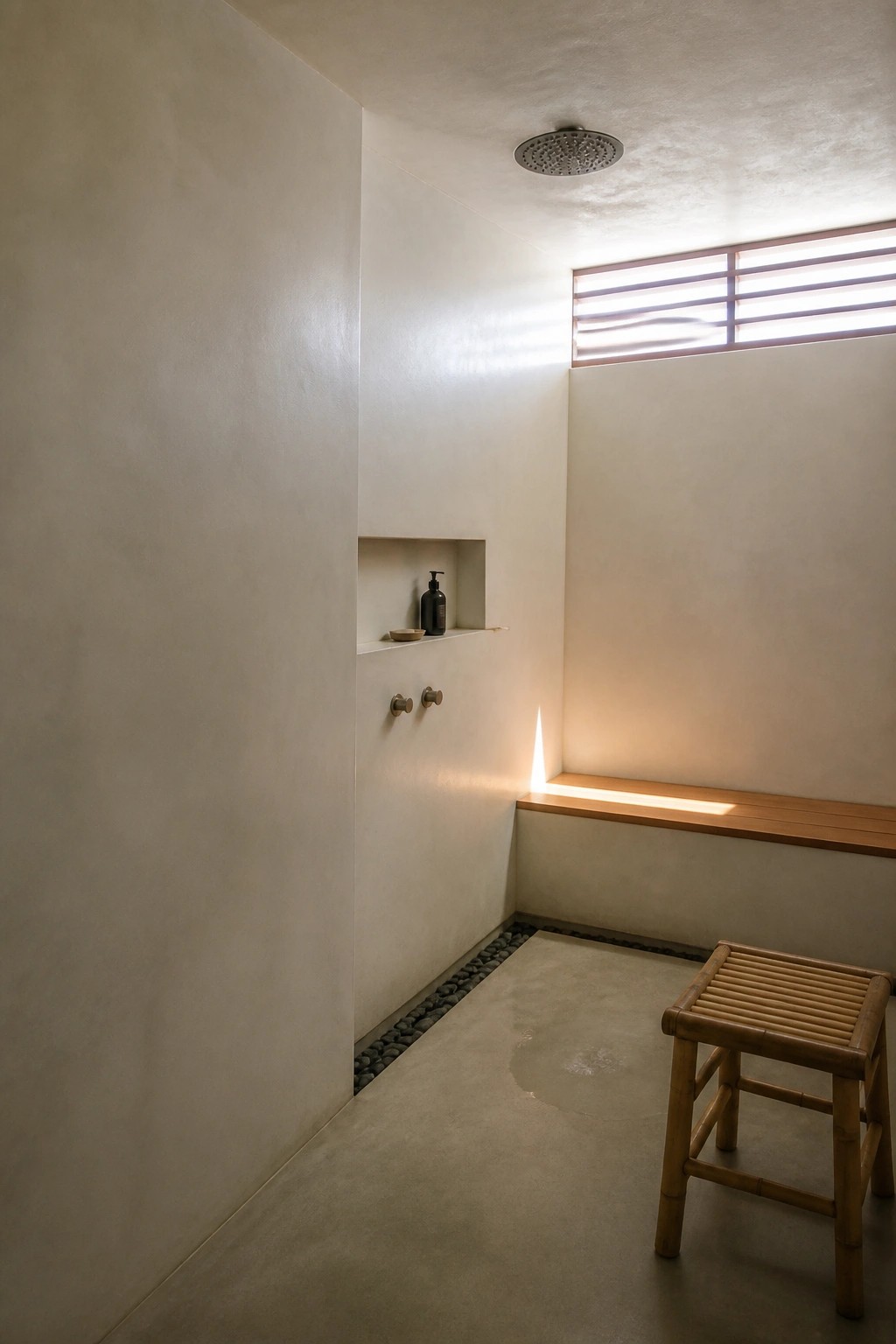 Minimalist shower room with plain walls, a ceiling mounted rain shower head, recessed niche with bottles, two small round wall controls, wooden bench, and pebble lined floor drain.