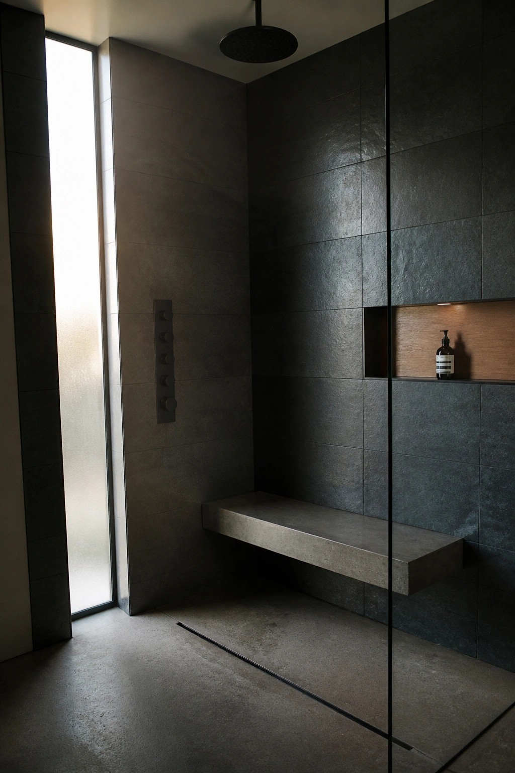 A minimalist dark tiled shower with a vertical flush control panel on the left wall, a floating concrete bench, and a recessed wooden niche holding one bottle.