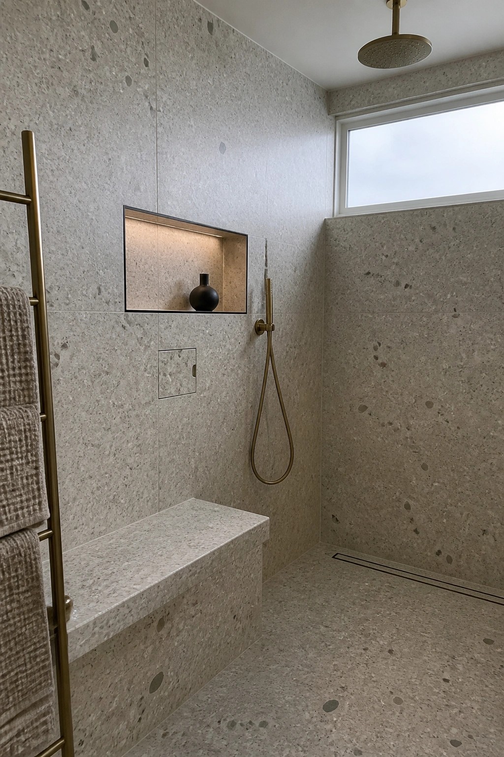 A minimalist shower interior with speckled beige tile, a recessed illuminated niche with a small black vase, brass rain head and handheld shower, built-in bench, towel rack with beige towels, and a small flush square access panel on the wall.