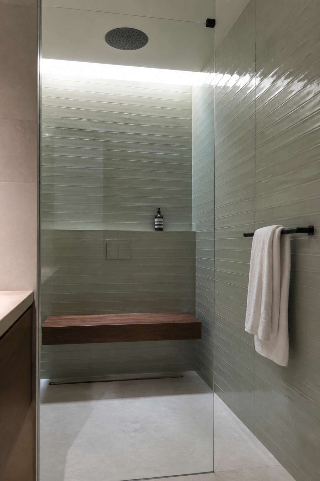 A modern walk-in shower with sage green horizontal tiles, a floating wood bench, ceiling-mounted rain showerhead, linear drain, and a white towel on a black bar outside the glass enclosure.