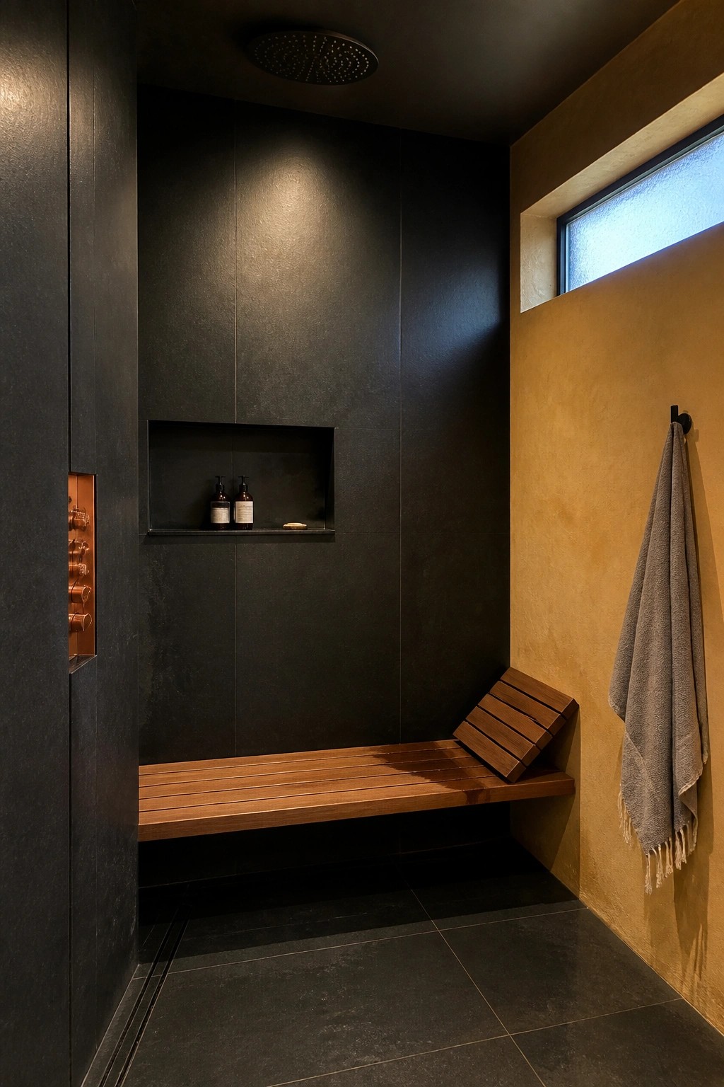 A minimalist shower with dark tiled walls, a built-in wooden bench, a recessed shelf holding bottles, and a flush-mounted copper control panel on the left wall.