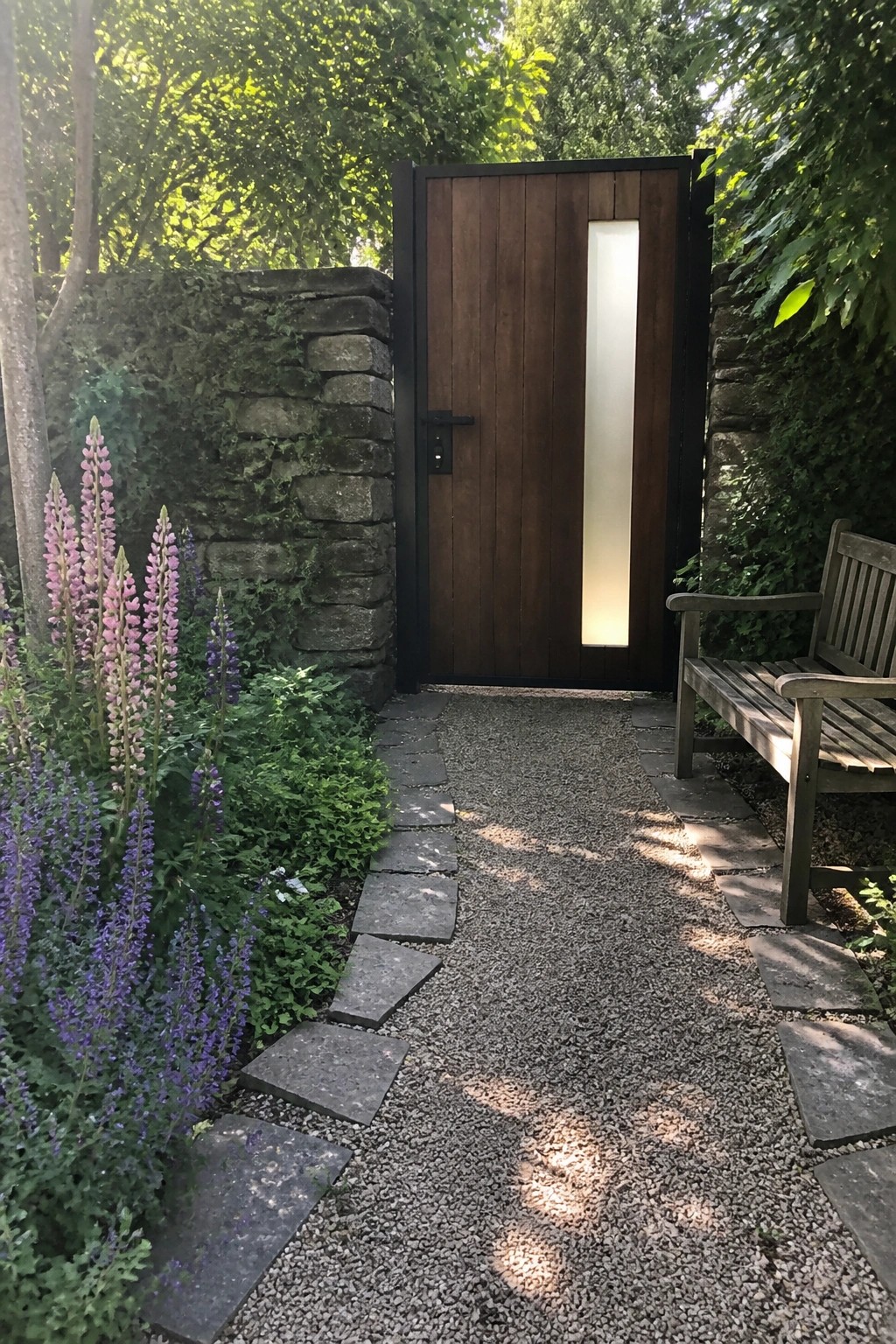 A wooden door with vertical frosted glass set in a black metal frame within a stone wall, bordered by a gravel path with stepping stones and flowering plants.