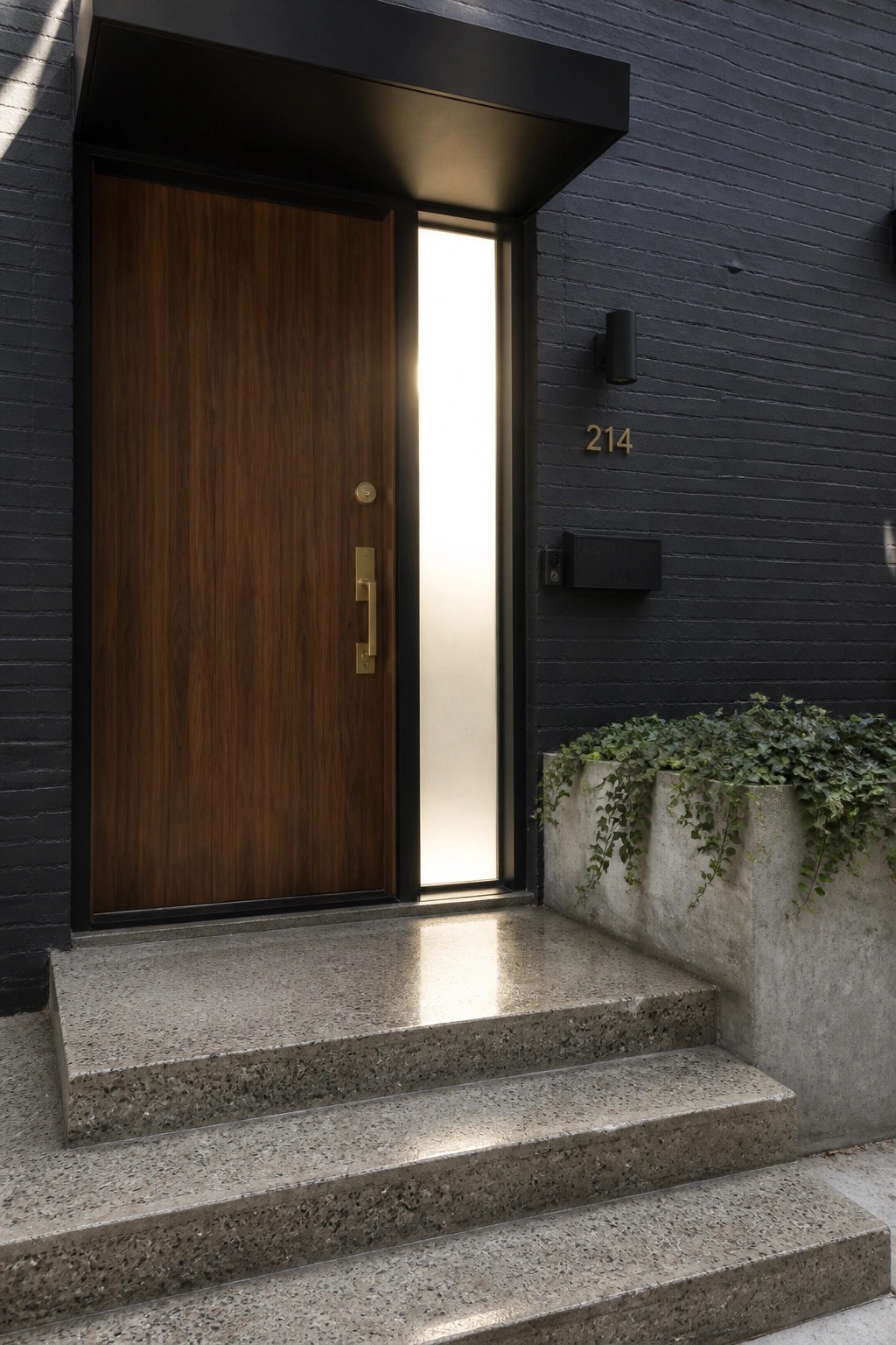A dark brick house exterior with a tall walnut wood front door, vertical frosted glass sidelight, concrete steps, and a small black mailbox mounted on the wall.