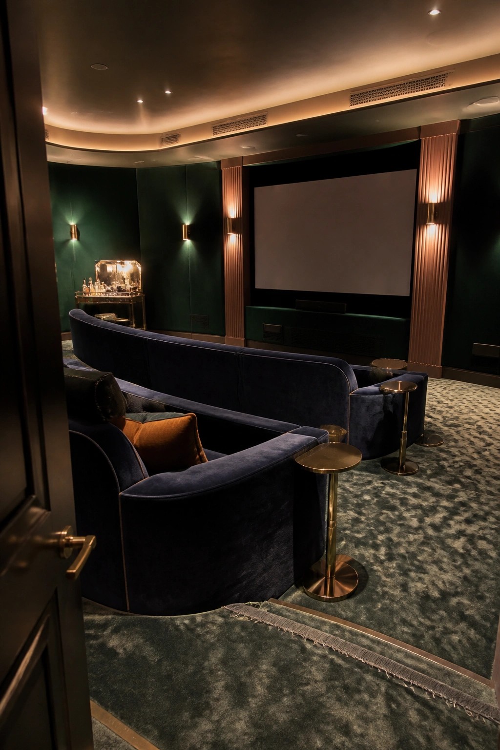 Interior view of a home cinema with curved dark blue velvet seating facing a large screen, set against green paneled walls with brass side tables.
