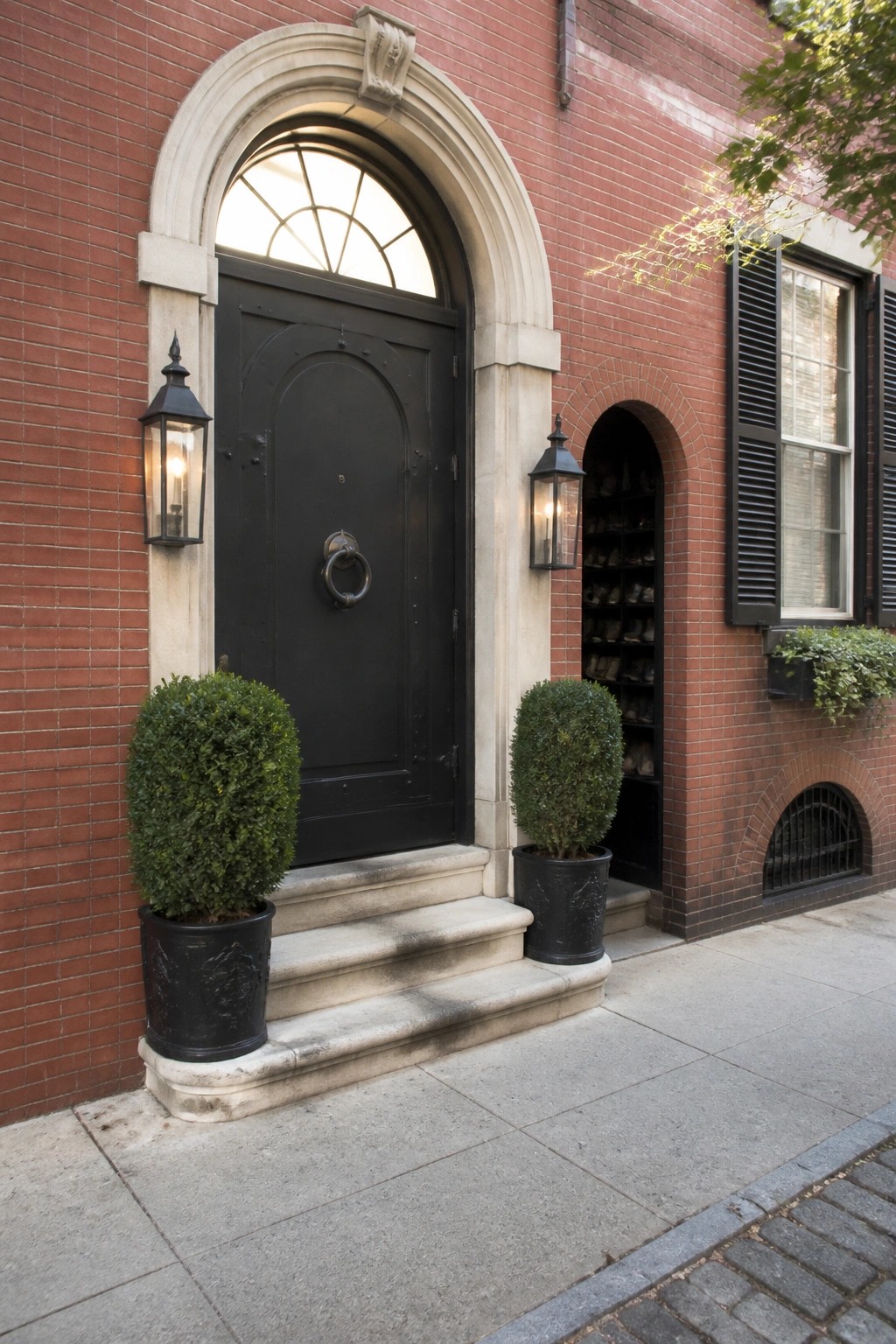 A large black arched door with a metal ring knocker is centered under a stone arch on a red brick building, with two potted shrubs and lanterns on either side.