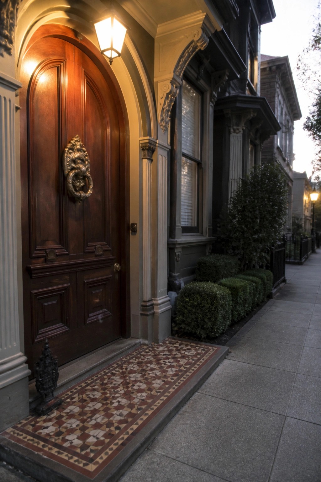 A large dark wood arched front door with an ornate metal knocker set into a painted house facade beside a tiled entry path.