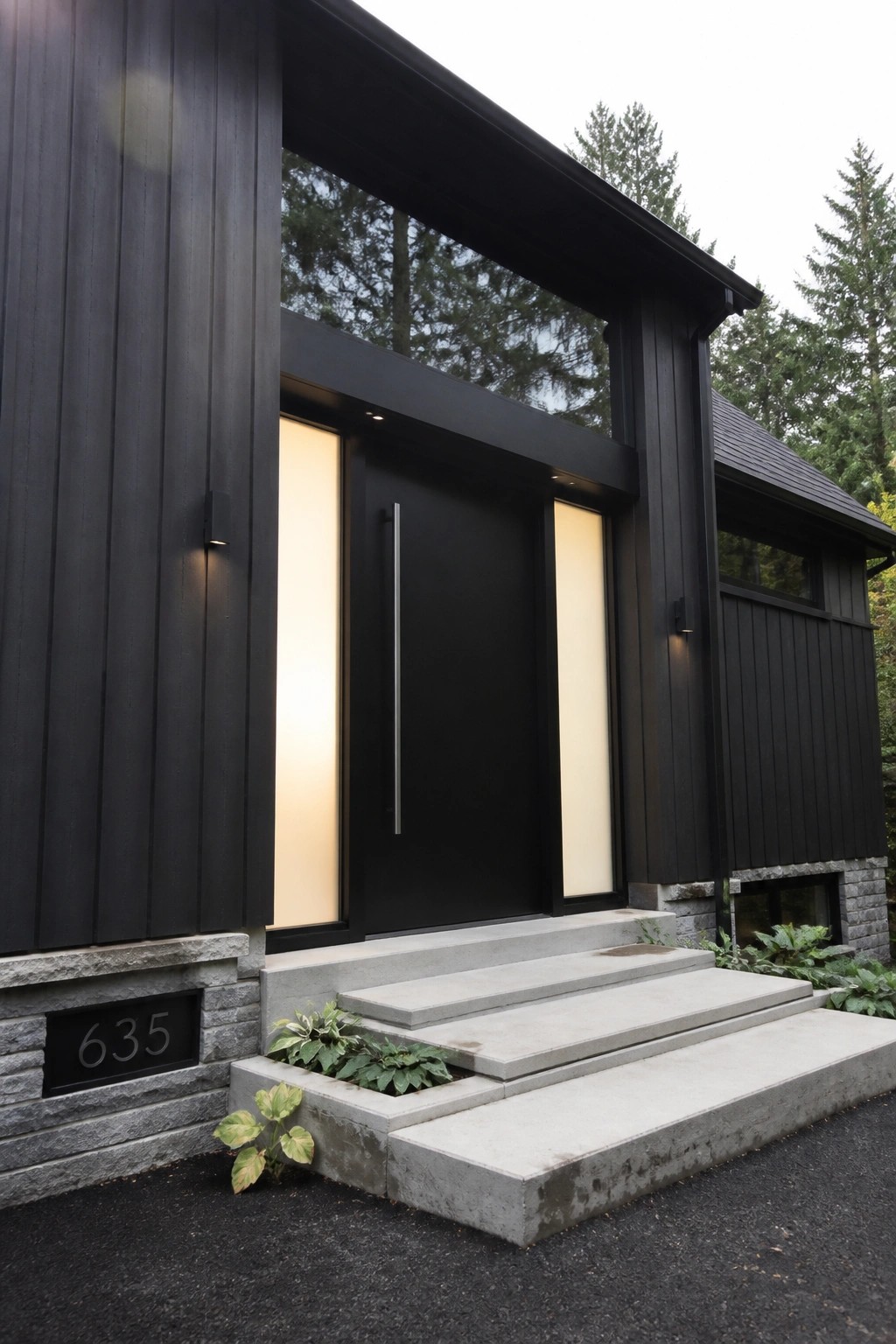 A modern black house exterior featuring a large black front door with a tall vertical metal handle, flanked by two tall frosted glass panels, concrete steps, and a house number plaque on a stone base.
