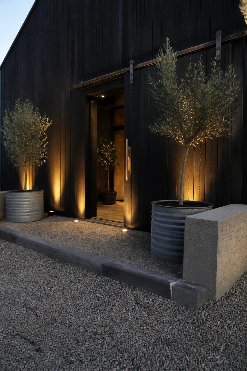 A black wooden exterior wall with a large open sliding door, metal handle, and two potted trees in corrugated metal containers on gravel.