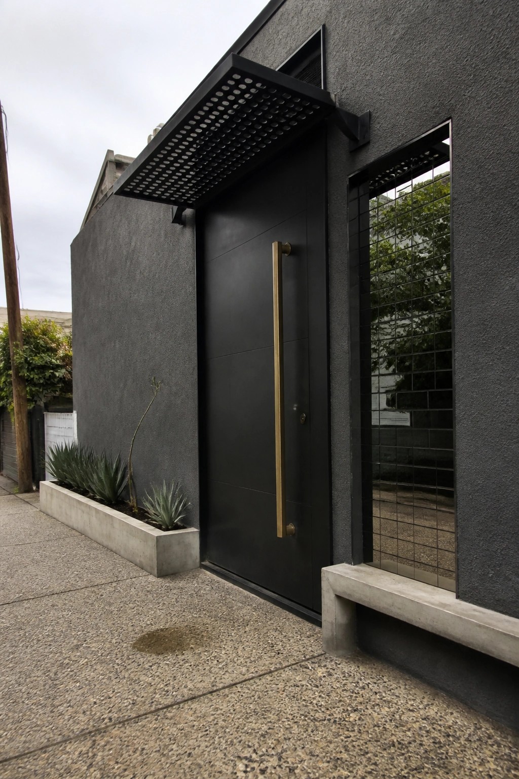 Modern black door with brass handle beside tiled window on gray building with concrete planters