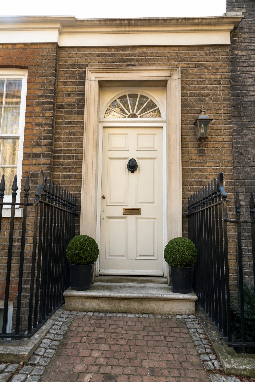 White door with fanlight on brick building, flanked by potted topiaries and iron railings.