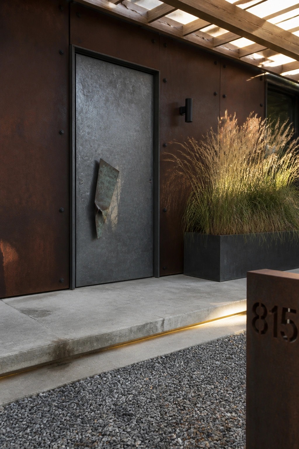 A dark metal door with an abstract sculptural accent mounted on a rusted steel facade next to a concrete planter with tall grasses.
