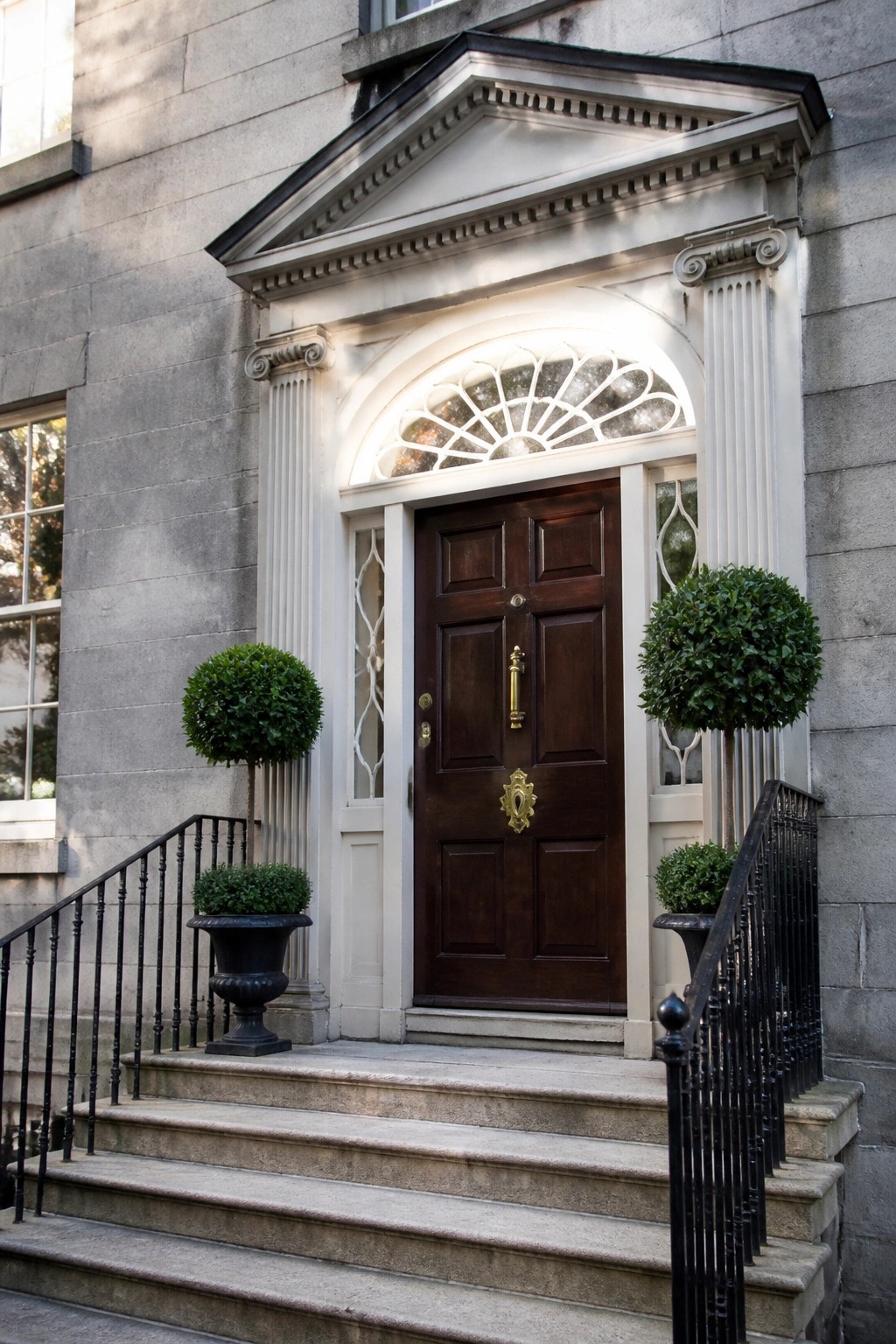 Front view of a dark wood double door with ornate brass hardware, set under a white pediment with an arched transom window on a stone building.