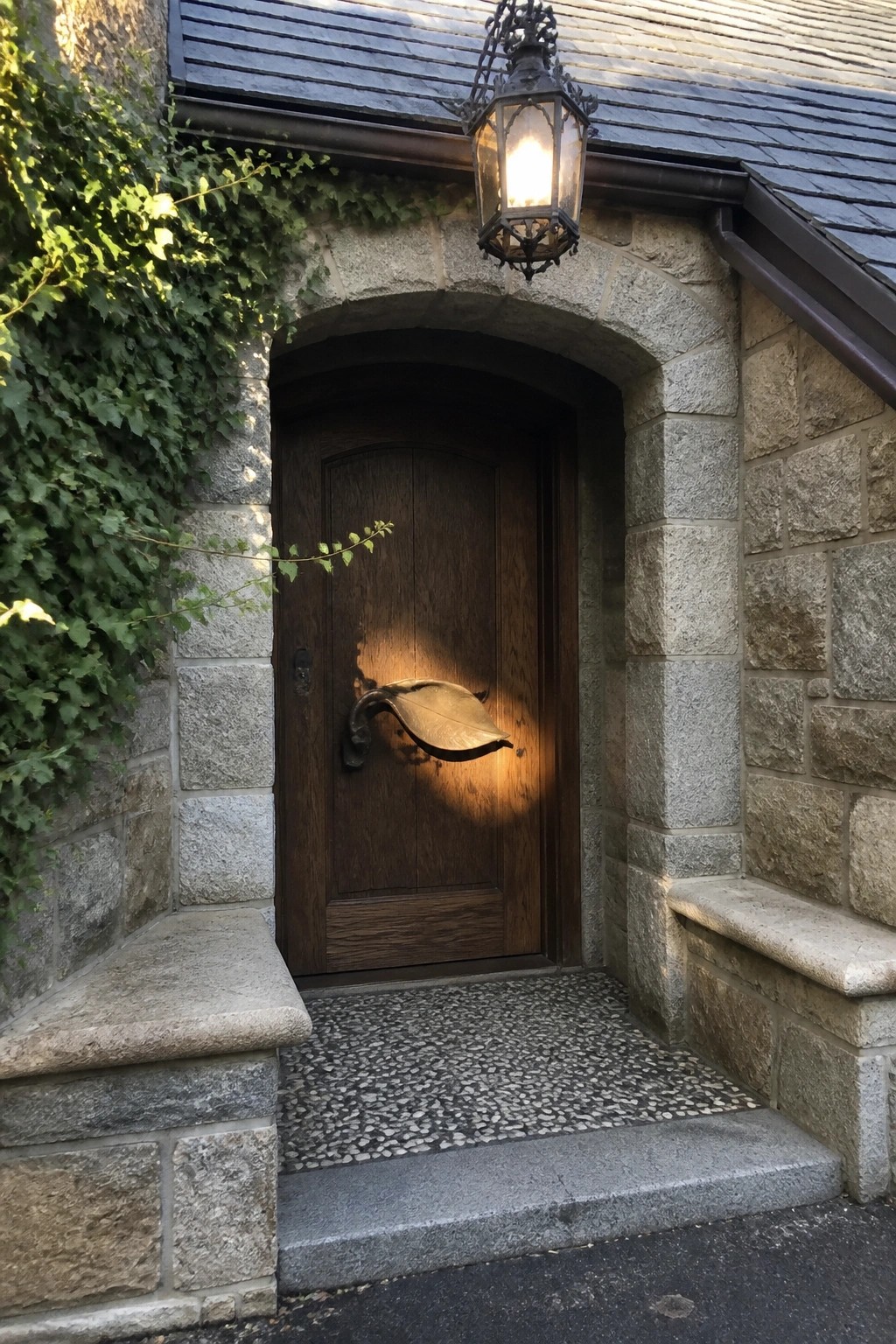 Wooden door with leaf knocker beneath glowing lantern in ivy-covered stone archway