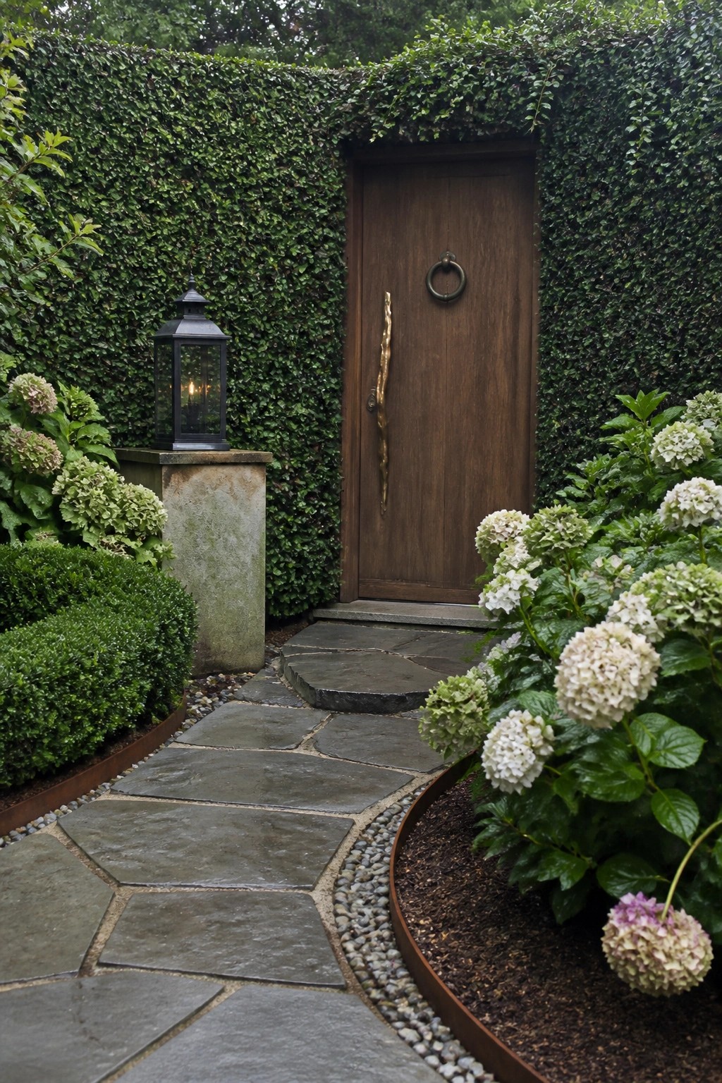 A wooden single door with a long sculptural handle and ring pull, set into a tall green hedge wall and reached by a curving stone path bordered by white hydrangeas and a black lantern.