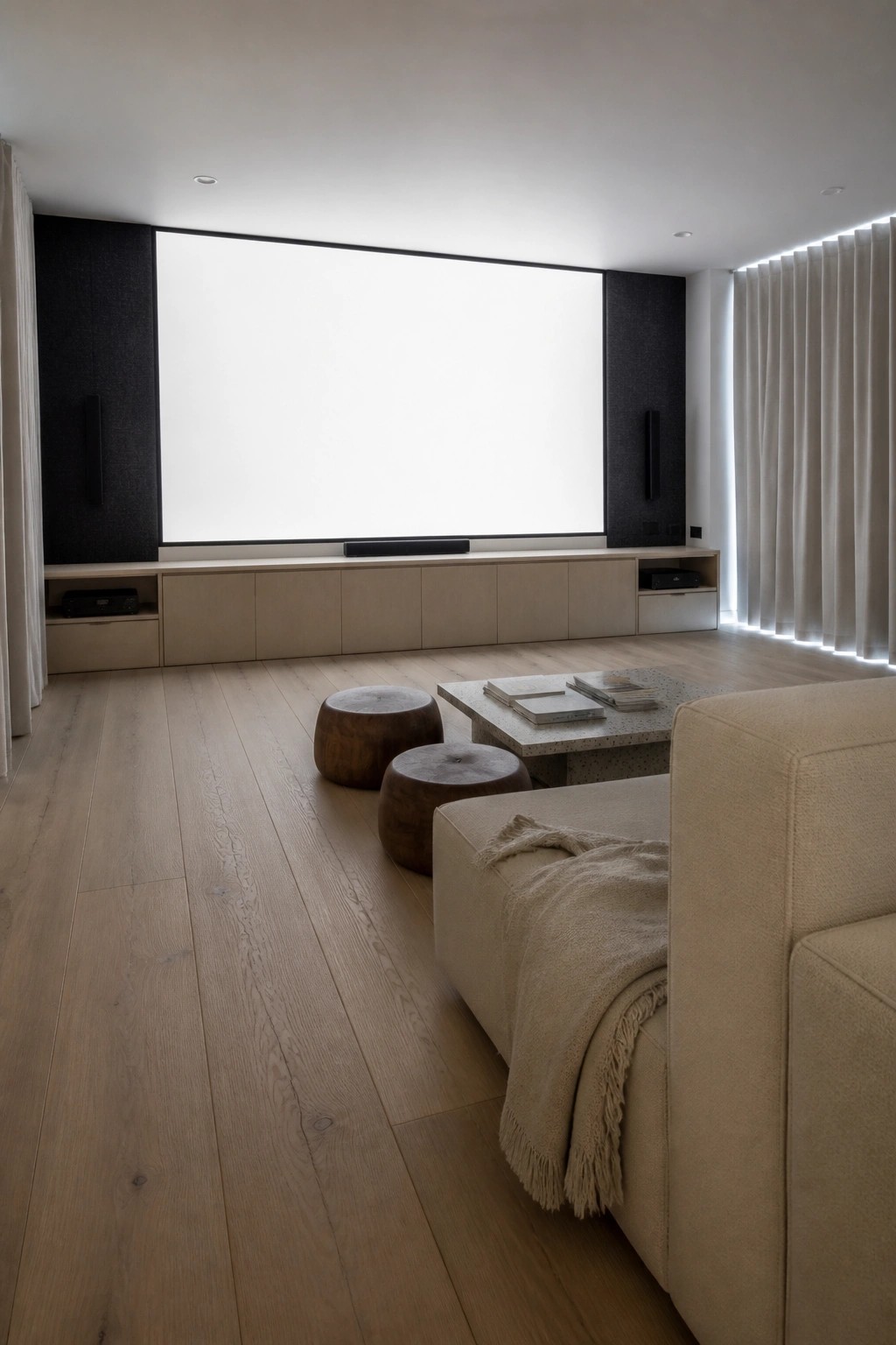 A minimalist interior room featuring a large wall-mounted screen above a continuous light wood cabinet, with two wooden stools and a sofa partially visible in the foreground.