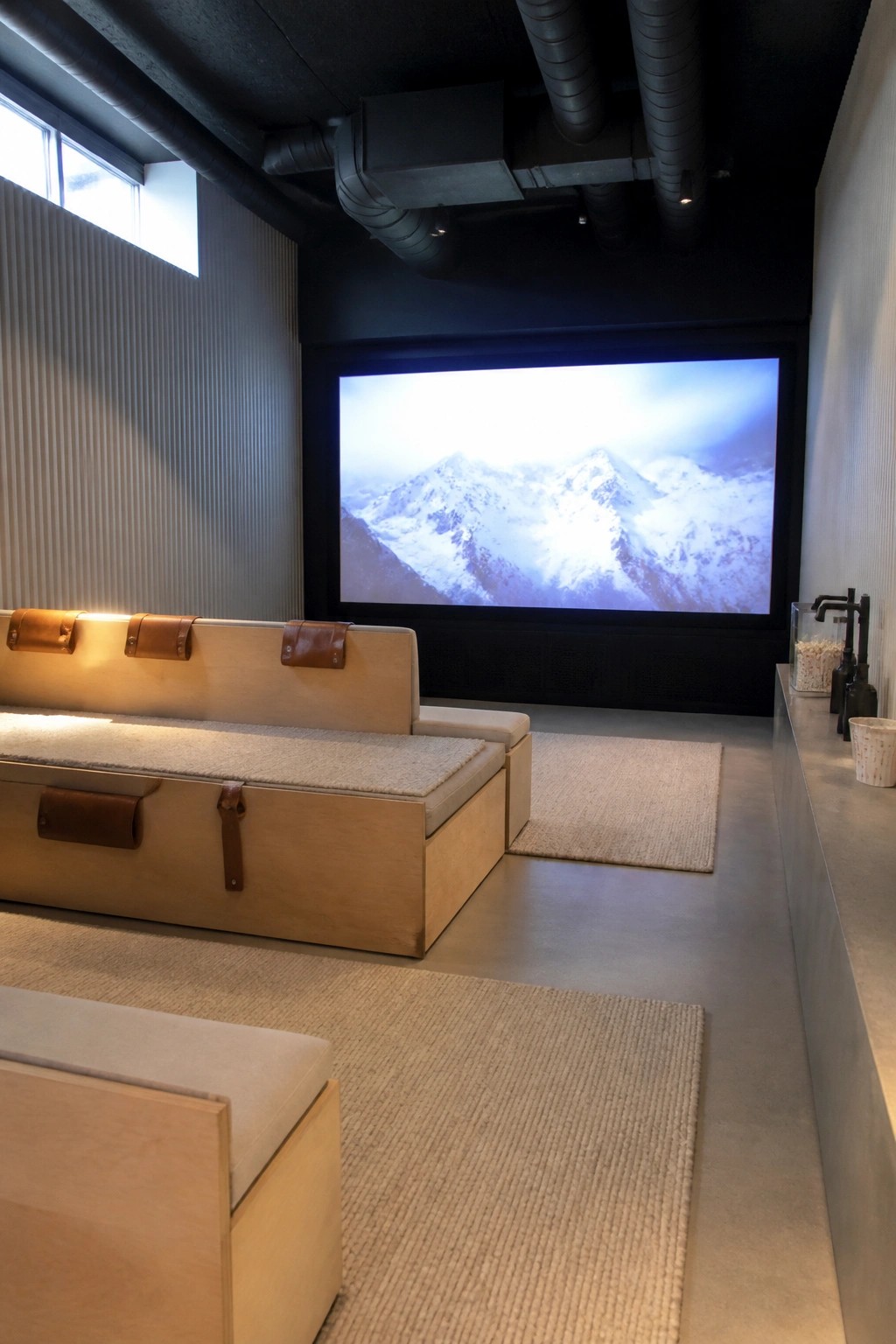 A modern home cinema room featuring a large floor-to-ceiling screen displaying snowy mountains, low modular wooden seating with beige cushions and brown leather straps, exposed black ceiling pipes, and light-colored rugs on a concrete floor.